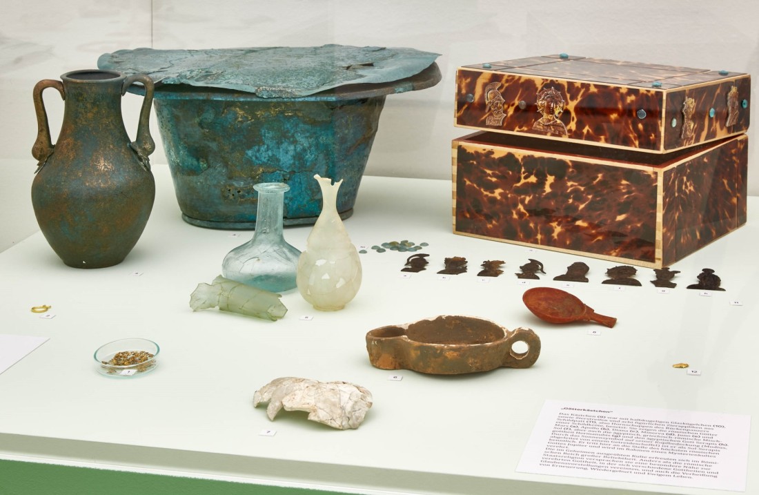 Funde aus Erkelenz-Borschemich, die einer Priesterin mit ins Grab gegeben wurden. Glasfläschchen, eine metallene Schüssel, ein Kästchen aus Schildpatt mit Götterfiguren davor.