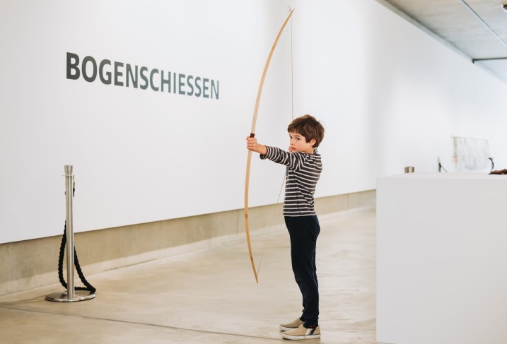 Ein Junge steht im Museum und hält einen großen BOgena us Holz in der Hand.