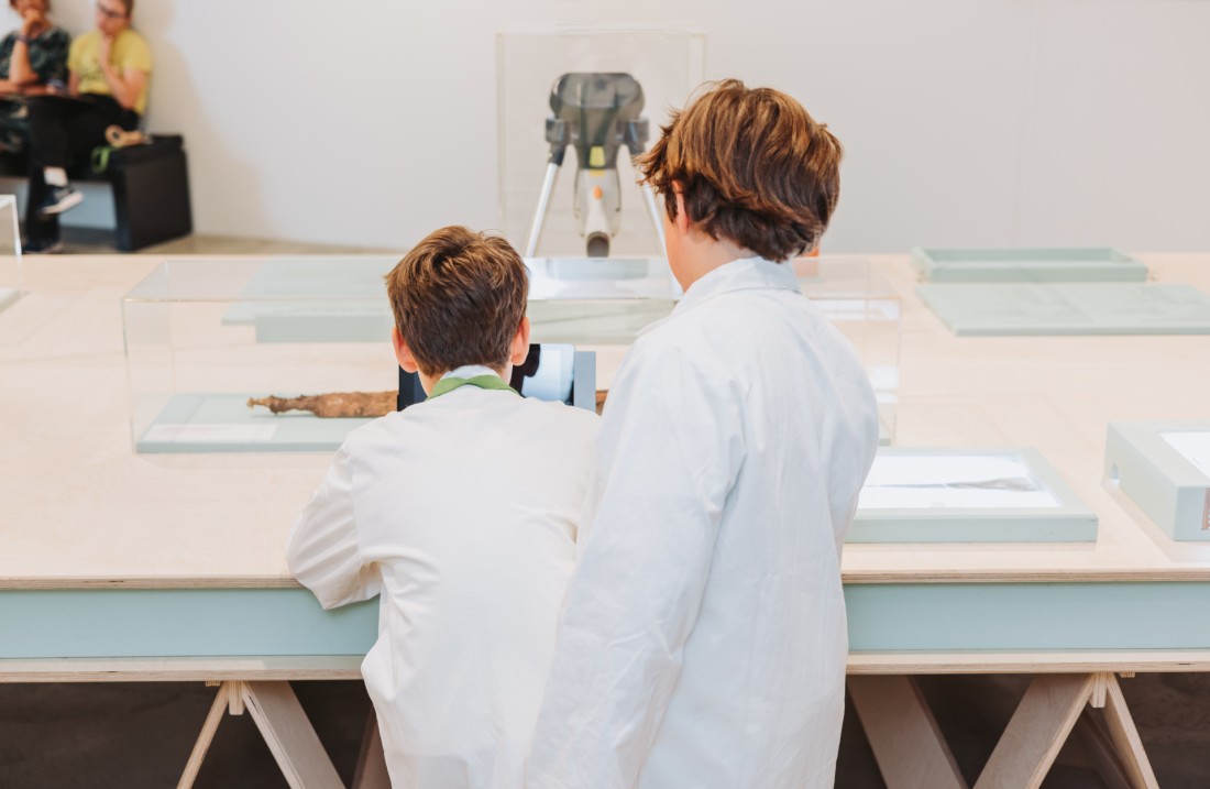 Zwei Jungen von hinten, die vor einem Arbeitstisch stehen. Foto: Katja Velmans © LVR-LandesMuseum Bonn 