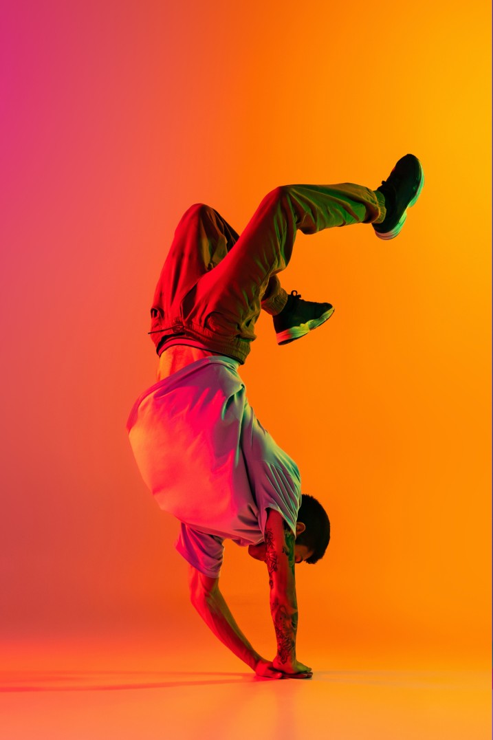 Vor einem orangefarbenen Hintergrund macht ein Tänzer in Sportkleidung einen dynamischen Handstand.