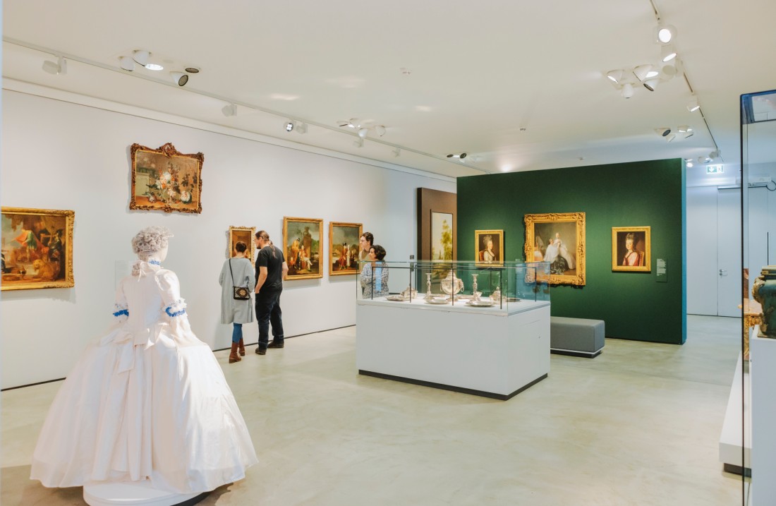 In einem geräumigen Ausstellungsraum hängen barocke Gemälde an den Wänden und eine Ankleidepuppe steht in der Raummitte. Einige Besucher und Besucherinnen schauen sich die Exponate an.