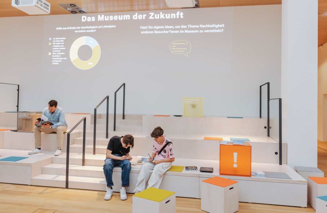 Auf einer Tribüne aus hellem Holz sitzen zwei Jugendliche und ein Mann, die jeweils vertieft etwas lesen oder schreiben. Im Hintergrund werden Informationen zum Museum der Zukunft an die Wand projeziert.
