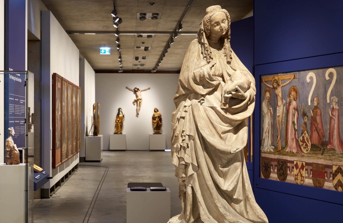 Eine Madonnenskulptur steht in einem langen Austellungeraum. Es sind weitere mittelalterliche Gemälde und Skulpturen zu sehen.