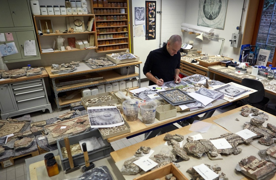 Ein Mann arbeitet an einem Tisch, der mit vielen kleinen Schachteln voll steht, in denen Teile des Mosaiks gelagert werden.