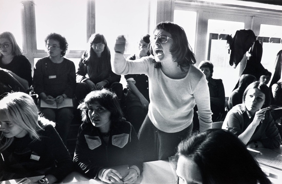 Eine Schwarz-Weiß-Fotografie von einem mit jungen Frauen gefüllten Innenraum. Eine der Frauen steht an einem Tisch und spricht und gestikuliert enthusiastisch.