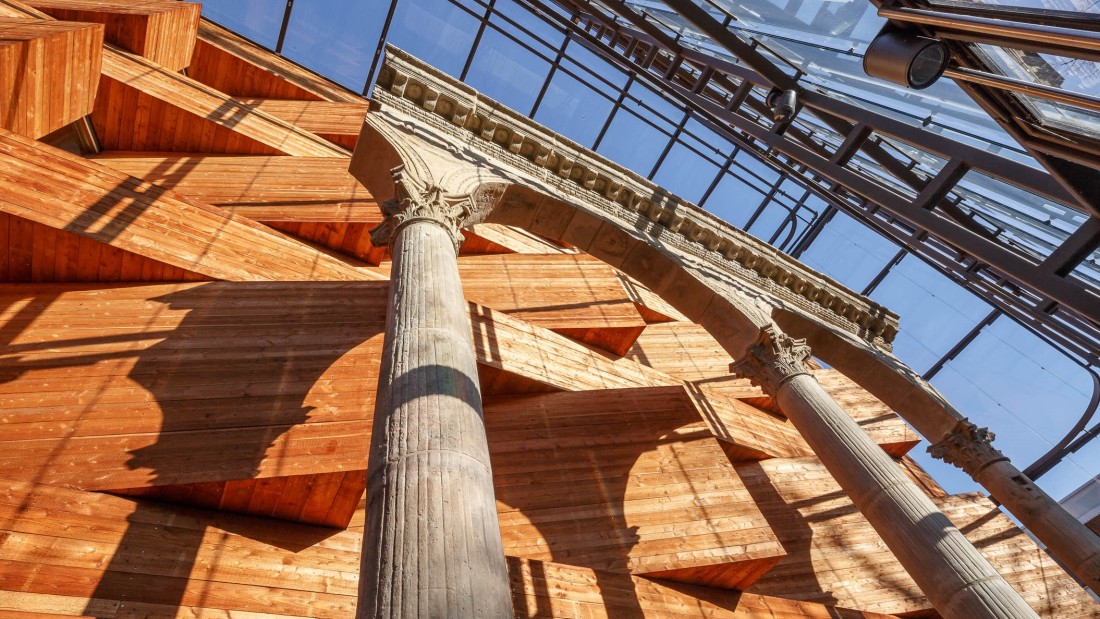 Blick an der Holzfassade des Museums entlang. Davor steht ein römischer Bogen. 