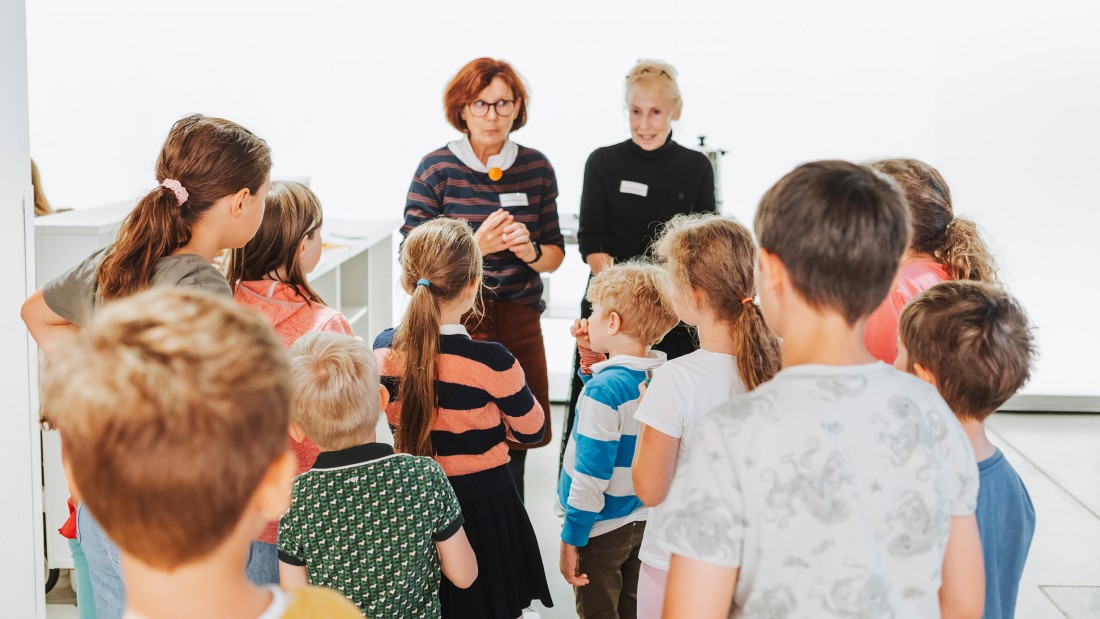 Eine Gruppe von Kindern hört den beiden Workshopleiterinnen aufmerksam zu. 