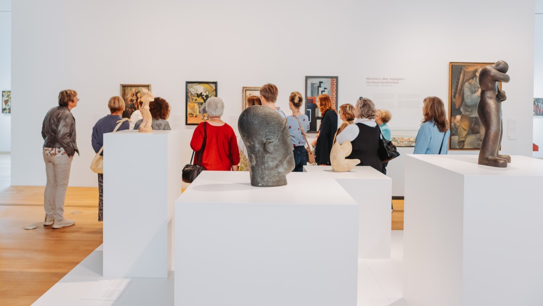 Eine Personengruppe wird durch die Ausstellung geführt. Zu sehen sind Bilder und Skulpturen. 