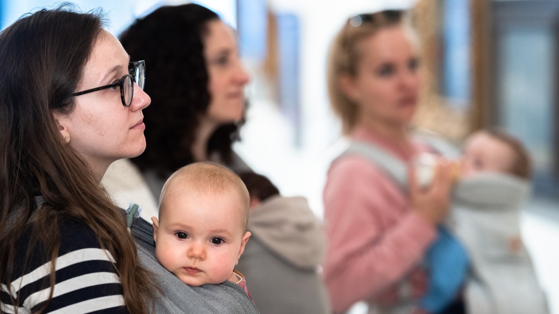 Frauen mit Babys in Tragen blicken auf ein Ausstellungsobjekt. 
