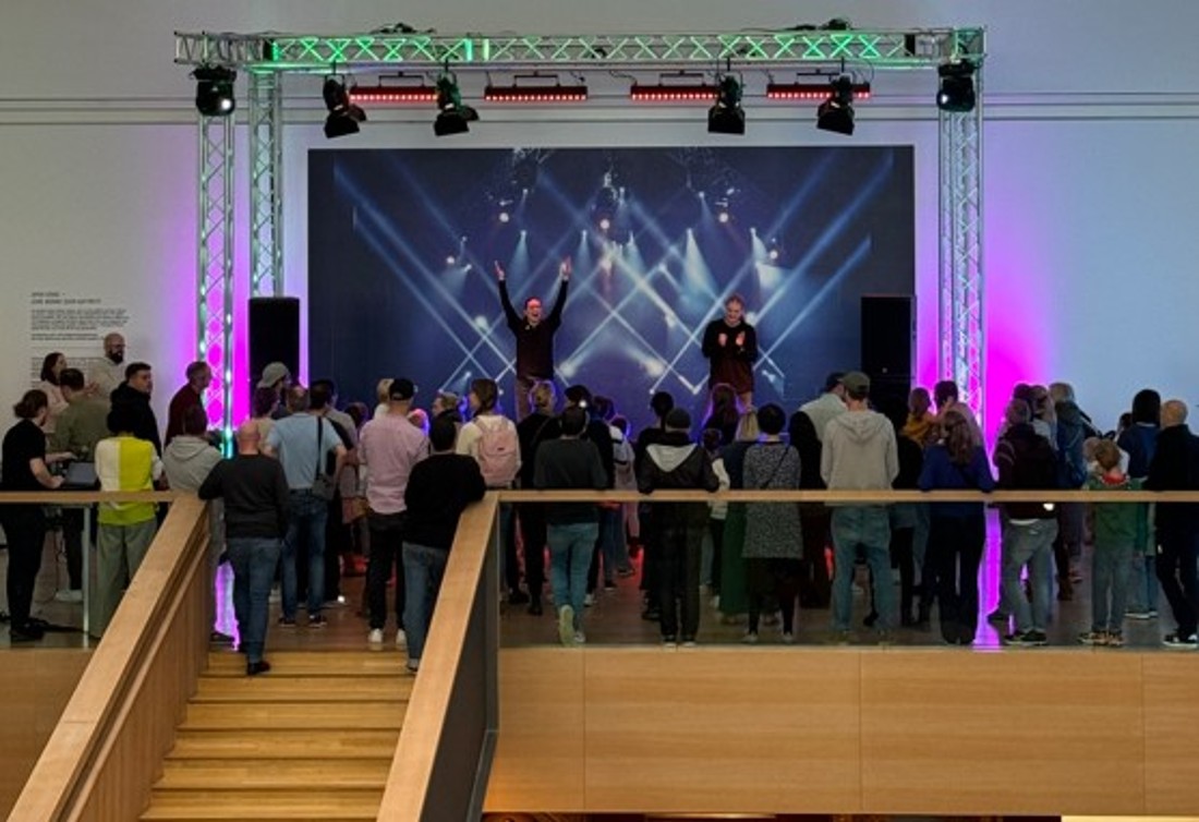 Menschen stehen vor einer B&uuml;hne im Museum. Auf der B&uuml;hne pr&auml;sentieren zwei K&uuml;nstler ihre Lieder.