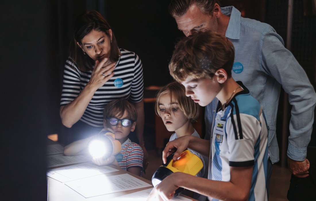 Eine Familie mit Taschenlampen ist &uuml;ber einen Tisch mit R&auml;tselfragen gebeugt.