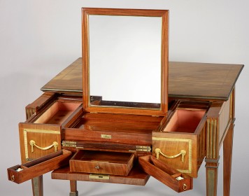 Der schlichte Verwandlungstisch besteht aus rötlichem Holz und goldenen Verzierungen. An den Seiten sind längliche Schubladen geöffnet und in der Mitte ein Spiegel hochgeklappt.