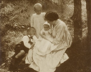 Im Mittelpunkt der Fotografie sitzt eine Mutter mit ihren drei Kindern auf einer Wiese. Rechts am Bildrand erkennt man den Stamm eines Baumes.