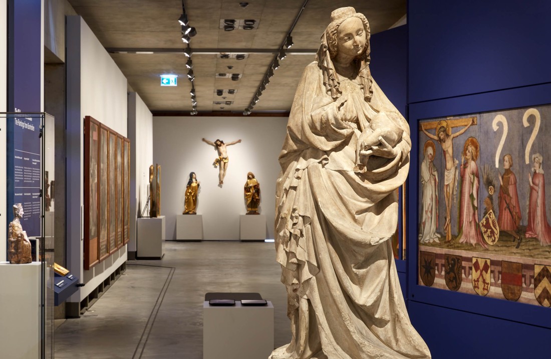 Ein lang gestreckter Ausstellungsraum mit blau gestrichenen Wänden und mittelalterlichen Gemälden. Im Vordergrund steht eine Marienskulptur und im Hintergrund hängt eine Kreuzigungsgruppe an der Wand.
