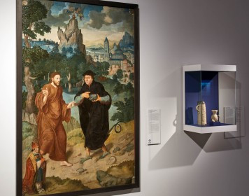 An einer Wand hängt ein Gemälde, auf dem der Teufel in Gestalt von Martin Luther Jesus in Versuchung führt. Daneben ist in einer Wandvitrine Steinzeug ausgestellt.