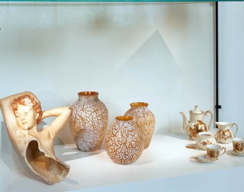 In einer Vitrine stehen diverse verzierte Alltagsgegenstände: Teetassen und eine Kanne, Vasen und eine zerbrochene Skulptur eines weiblichen Oberkörpers.