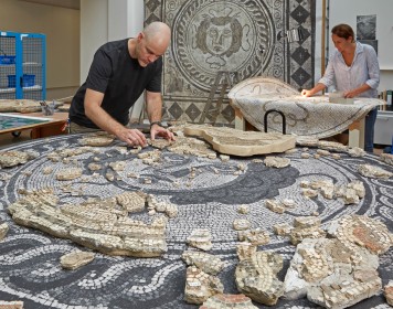 An einem großen Tisch arbeitet ein Restaurator an den Steinen eines römischen Mosaiks.