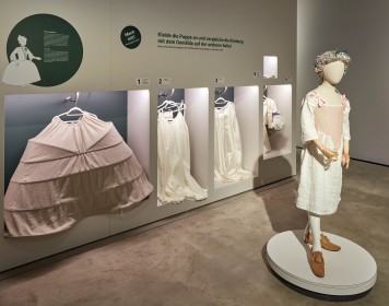 Eine lebensgroße Kleiderpuppe steht im Vordergrund im Ausstellungsraum. Hinter ihr hängen verschiedene barocke Kleidungsstücke.