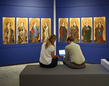 Vor den Heiligenfiguren des Bornhofener Retabels sitzen zwei Personen auf einer Bank im Ausstellungsraum und schauen gemeinsam auf ein Tablet.