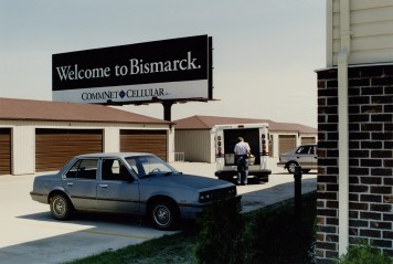 Eine Farbfotografie, auf der ein Auto und einfache Häuser zu sehen sind. Auf einem Schild steht Welcome to Bismarck.