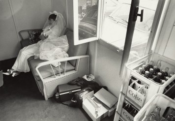 Eine Frau im Brautkleid sitzt mit verschränkten Armen neben einem geöffneten Fenster, danaben Cola-Kästen.