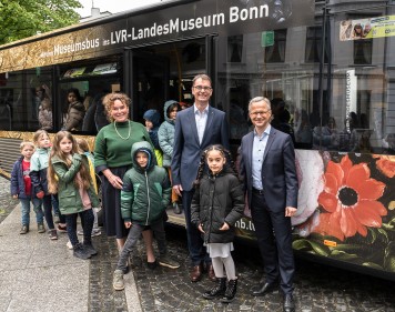 Im Hintergrund der Museumsbus, vor der Bustür die drei Partner Stadtsparkasse, SWB und LVR-Landesmuseum Bonn, davor Kinder.