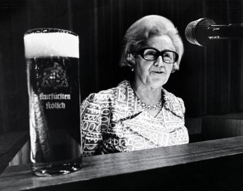 Die Schwarz-weiß-Aufnahem einer Frau, die an einem Rednerpult steht – vor ihr steht prominent ein gefülltes Kölschglas.