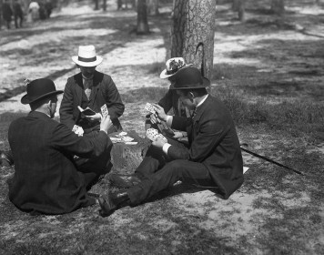 Die Schwarz-weiß-Fotografie zeigt vier Männer mit Hüten, die in einem Park auf dem Boden sitzend Skat spielen.