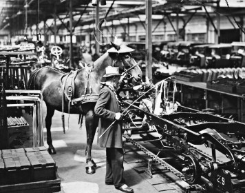 Eine schwarz-weiße Fotografie, die einen Droschkenkutscher mit Pferd am Fließband des Opelwerks in Rüsselsheim zeigt.
