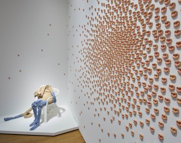 Viele einzelne Kussmünder an eine weiße Wand geklabt. Im Hintergrund eine kopflose Figur auf einem Gartenstuhl sitzend.